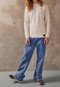 White, ribbed long-sleeve top with a small black logo, paired with loose-fitting blue jeans and dark athletic shoes. Simple and casual design.