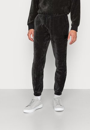 Black velour joggers with elastic cuffs, drawstring waist, and a textured surface. Worn with light grey sneakers and white ankle socks.