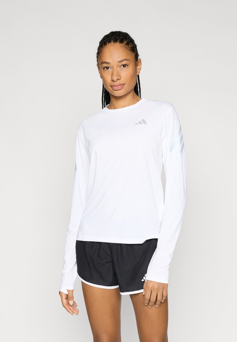 Camiseta deportiva blanca de manga larga con detalles reflectantes, combinada con pantalones cortos negros con ribete blanco y logo, adecuada para actividades deportivas.