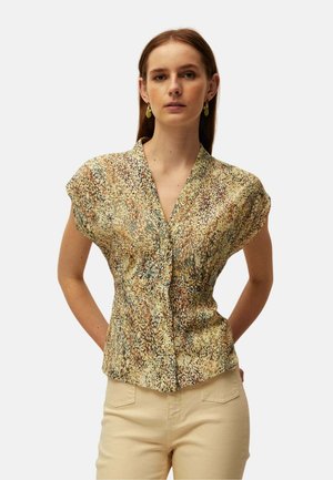 Mujer con cabello castaño liso lleva una blusa de manga corta con estampado en tonos tierra y pantalones beige, de pie con las manos detrás de la espalda.