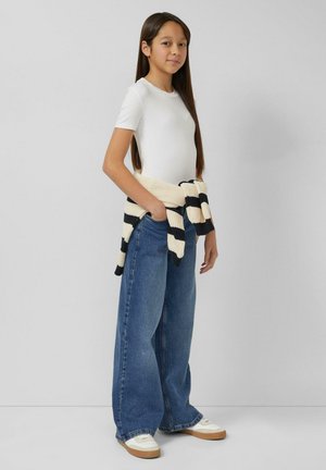 Mädchen mit langen braunen Haaren, das ein weißes T-Shirt, weite blaue Jeans, weiße Turnschuhe und einen gestreiften Pullover, der um die Taille gebunden ist, trägt und seitlich steht.