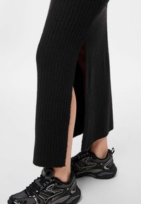 Črna rebrasta pletena obleka z bočnimi razporki, v kombinaciji s črnimi športnimi čevlji z mrežastimi in sintetičnimi detajli.