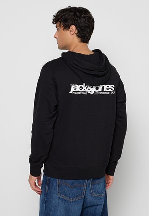Muž otočený bokom, oblečený v čiernej mikine s kapucňou a bielym logom "jack & jones project core" na chrbte a modrých džínsach na jednofarebnom pozadí.