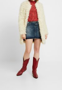 Cappotto in pelliccia sintetica color crema, top rosso con paillettes, minigonna in denim blu e stivali da cowboy rossi in pelle con punta appuntita e bordi scallop.