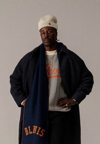 Musta takki, harmaa neule punaisilla kirjaimilla, sininen huivi oransseilla yksityiskohdilla, beige pipo. Asusteina kerroksittain kaulakoruja ja sormuksia.