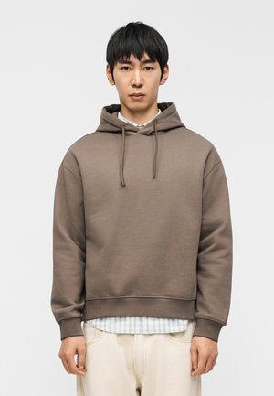 Brown hooded sweatshirt with drawstrings, ribbed cuffs and hem, layered over a blue checkered shirt, worn with light-colored pants.