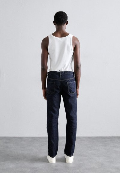 Dunkelblaue Jeans mit dezenten Nähten, kombiniert mit einem weißen Tanktop. Das Modell ist von hinten zu sehen und zeigt eine elegante gerade Passform.