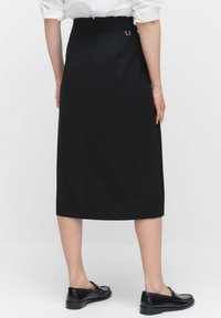 Vrouw in een zwarte knielange rok met een rits aan de achterkant, een witte blouse en zwarte leren loafers, staande tegen een effen achtergrond.