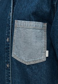 Τζιν μπουφάν με σκούρο μπλε ύφασμα, αντίθετη τσέπη σε ανοιχτό μπλε και καφέ ραφές. Ορατές λεπτομέρειες κουμπιών στο μπροστινό μέρος.