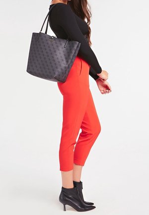 Femme portant un pantalon rouge court, un haut noir à manches longues, des bottines noires, portant un grand sac cabas à motifs sur l'épaule.