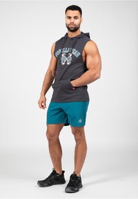 Mouwloze donkergrijze hoodie met grafische tekst, gecombineerd met teal shorts. Model draagt zwarte sportschoenen en toont een gespierd postuur.