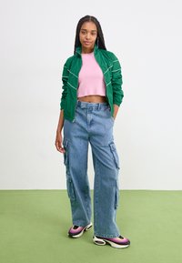 Green zip-up jacket with white stripes, pink cropped top, blue cargo jeans, and multicolored sneakers, set against a plain background.