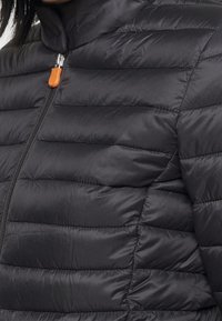 Sort pufferjakke med et quiltet design, der har vandrette syninger og en høj krave. Den har en sølvfarvet lynlås og en orange trækstrop.