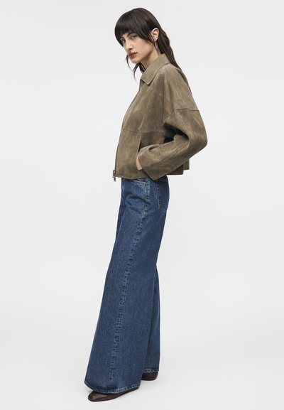 Žena v olivovo zelenej krátkej semišovej bunde, širokých modrých džínsach a hnedých topánkach, stojaca v profile s rukami v vreckách bundy.