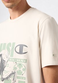 Mężczyzna w off-white koszulce z dużą grafiką zielonej i szarej modliszki oraz abstrakcyjnym tekstem na klatce piersiowej, a także małą naszywką z logo na rękawie.