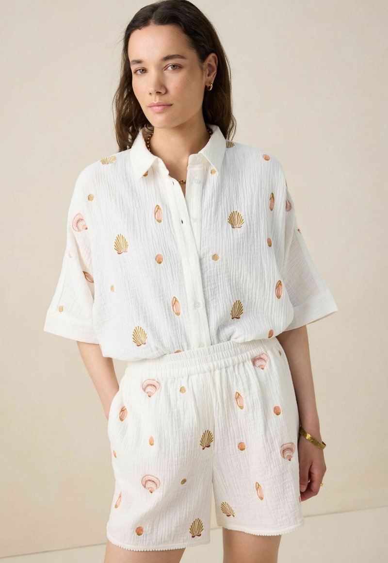 Ensemble en coton blanc comprenant une chemise à boutons et un short, tous deux brodés de motifs de coquillages roses et dorés. Tissu doux et texturé.