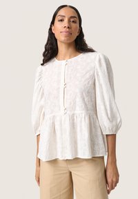 Blusa bianca con motivo floreale testurizzato, colletto rotondo, maniche a sbuffo e chiusura con bottoni. Vestibilità ampia con orlo svasato.