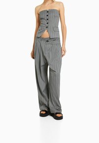 Kobieta ubrana w szary, prążkowany, bezramiękowy top zapinany na guziki oraz pasujące szerokie spodnie, do których nosi czarne sandały na platformie.
