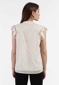 Blusa beige a maniche corte con spalle arricciate, caratterizzata da un motivo testurizzato di piccole punti in rilievo e un orlo inferiore trasparente.