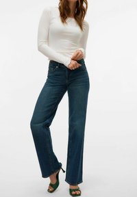 Bela dolgosleeve majica z ribbed teksturo, v kombinaciji s temno modrimi ravnimi hlačami. Hlače imajo visok pas in rahlo strgan rob.