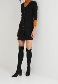 Musta napillinen mekko pitkillä hihoilla, yhdistettynä mustiin reiteen asti ulottuviin sukkiin ja korkokengillä. Sujuva rakenne ja istuva siluetti.