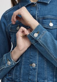 Mėlynas džinsinis švarkelis su sidabriniais mygtukais, dviem krūtinės kišenėmis ir mygtukais apvadais ant rankogalių. Audinys turi klasikinį, tekstūruotą išvaizdą.