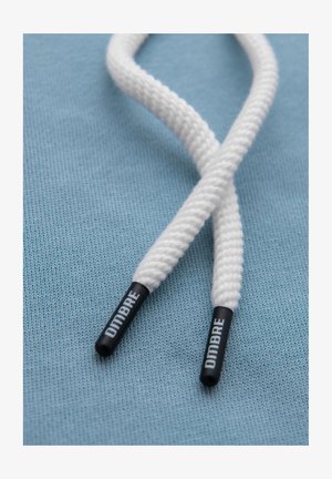 Białe sznurowadła o teksturowanym wykończeniu, z czarnymi, okrągłymi końcówkami (agletami) z napisem "DIMBRE", leżące na jasnoniebieskim tle tkaniny.