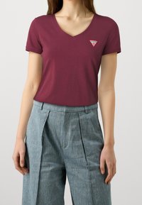 Burgundin värinen V-kaula-aukkoinen t-paita, joka on valmistettu puuvillasta ja jossa on pieni punainen ja valkoinen kolmiomainen logopatchi vasemmassa rinnassa.