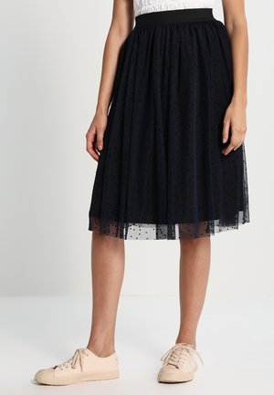 Mujer con falda midi negra de lunares con una sobreposición translúcida y zapatillas beige, de pie contra un fondo claro y liso.