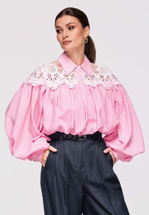 Kobieta nosząca różowo-białą bluzkę w paski z białym koronkowym karczkiem i bufiastymi rękawami, połączoną z ciemnymi spodniami z wysokim stanem.