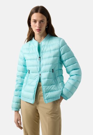 Kobieta w jasnoniebieskiej pikowanej kurtce z kieszeniami na zamek i beżowych spodniach, stoi z jedną ręką w kieszeni na białym tle.