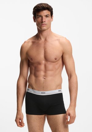 Muskelöser Mann steht und trägt schwarze BOSS Boxershorts mit weißem Bund vor einem einfarbigen weißen Hintergrund.