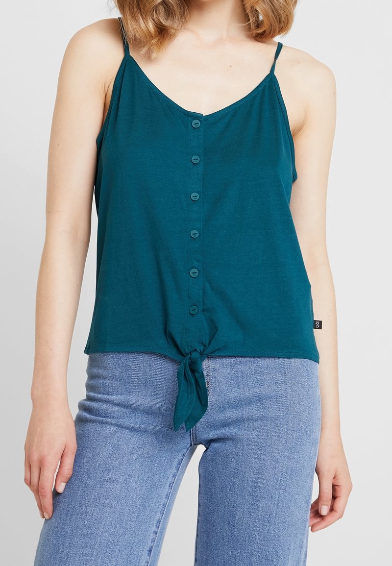 Turkos ärmlös topp med v-ringning, knapplist framtill och knuten nederkant. Mjuk tyg med avslappnad passform, i kombination med ljusblå jeans.