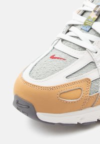 Αθλητικό παπούτσι Nike με γκρι υφασμάτινο επάνω μέρος, λευκές δερμάτινες λεπτομέρειες, καφέ λαστιχένιο κάλυμμα στα δάχτυλα και μικρό κόκκινο λογότυπο. Υφή με διακριτό φινίρισμα και αντίθετα υλικά.