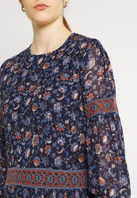 Marinblommig klänning med långa puffärmar, rund halsringning och intrikat broderi i orange och blått runt midjan och ärmarna.