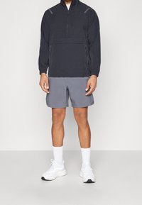 Zwarte zip-up jas met lange mouwen, grijze sportshorts en witte sportschoenen. Kenmerken zijn onder andere zijzakken en contrasterende accenten.