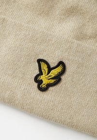 Beige kötött sapi puha textúrával, elöl fekete szegéllyel és sárga hímzett saslogóval. Egyszerű és funkcionális dizájn.
