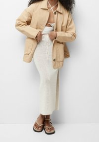 Beige katoenen jack, witte opengehaakte rok en tan sandalen met een band. Het jack heeft zakken en een losse pasvorm.
