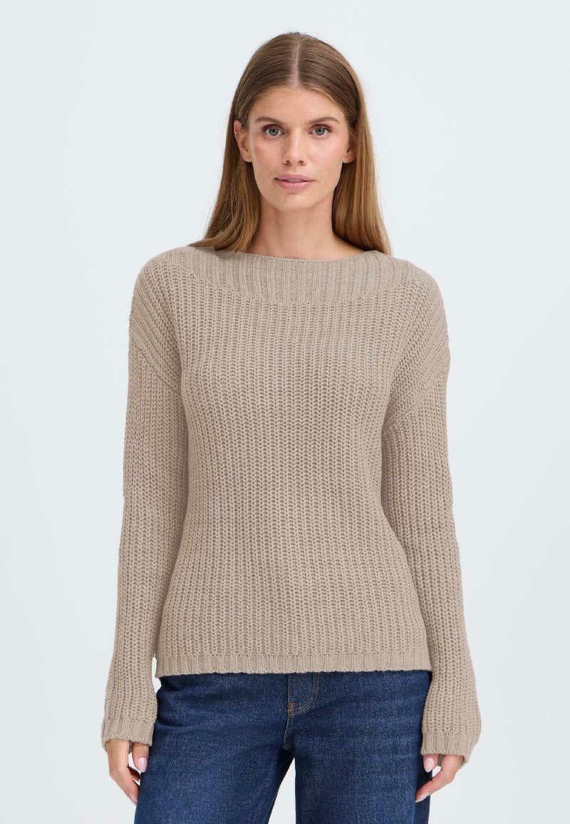 Beige strikkegenser med bred rund hals, lange ermer og ribbestrikket tekstur. Kombinert med mørkeblå jeans med avslappet passform.