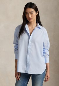 Kvinne med langt mørkt hår iført en lyseblå oversized skjorte og blå jeans, står mot en nøytral bakgrunn.