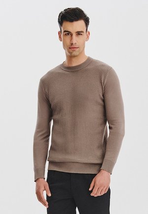 Brązowy sweter z dzianiny z długimi rękawami, fakturowanym przodem i ściągaczami przy mankietach. Noszony z czarnymi spodniami, ma okrągły dekolt.