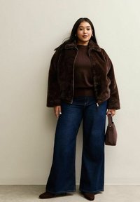 Donna che indossa un giaccone in pelliccia sintetica marrone scuro, un maglione abbinato, jeans a gamba larga blu scuro, stivali marroni e tiene una borsa marrone.