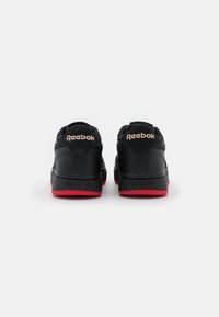Baskets Reebok noires avec une texture lisse, dotées de semelles rouges et d'une forme arrondie. Logo doré en accent à l'arrière.