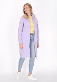 Jasnofioletowy długi płaszcz przeciwdeszczowy z zamkiem z przodu, z dwiema przednimi kieszeniami, noszony na żółtej bluzce w paski i jasnoniebieskich dżinsach, zestawiony z białymi trampkami.