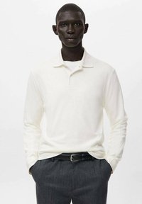 Langärmliges Poloshirt in Off-White, aus weichem Stoff, mit spitzem Kragen und schmaler Passform, kombiniert mit dunklen Nadelstreifenhosen.