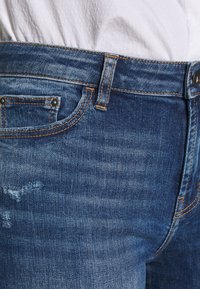 Jeans de mezclilla azul oscuro con un diseño clásico de cinco bolsillos, que presentan detalles de costura, cierre de botón y efectos de desgaste sutiles.