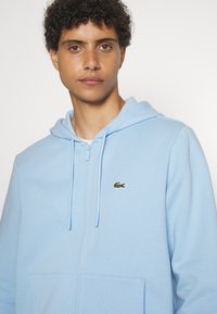 Šviesiai mėlynas užtrauktukas su kapučinu, pagamintas iš minkštos medžiagos. Yra kengūrinė kišenė ir mažas žalias „Lacoste“ krokodilo logotipas siuvinėtas ant krūtinės.