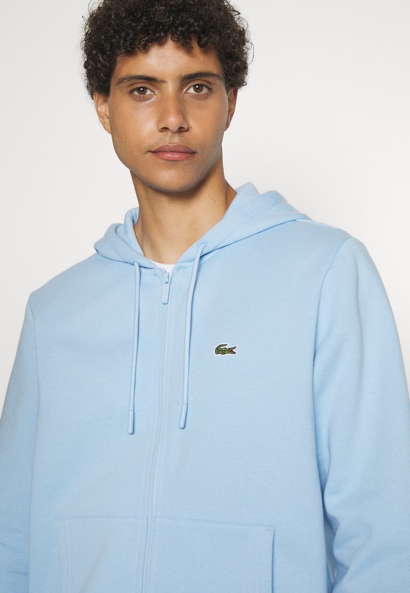 Helle blaue Zip-Hoodie aus weichem Stoff. Mit einer Kängurutasche und einem kleinen grünen Lacoste-Krokodil-Logo, das auf der Brust gestickt ist.
