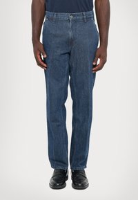 Hombre que lleva jeans rectos de color azul oscuro y mocasines de cuero negro, de pie contra un fondo claro y neutro.