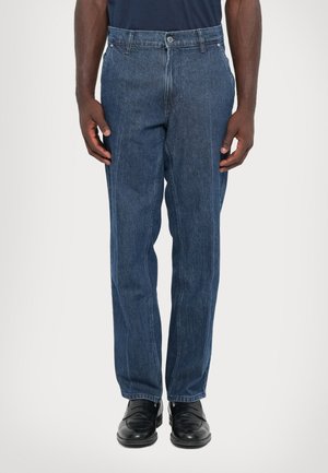 Man wearing dark blue straight-leg jeans and black leather loafers, standing against a plain light background.